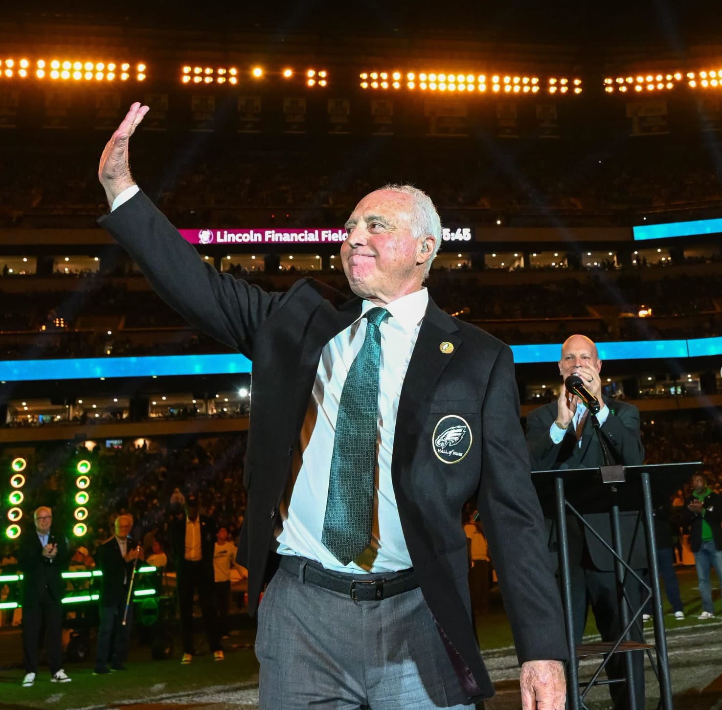 Jeffrey Lurie in a dark blazer with an Eagles Hall of Fame patch and a green tie, waving to the crowd on the field at Lincoln Financial Field